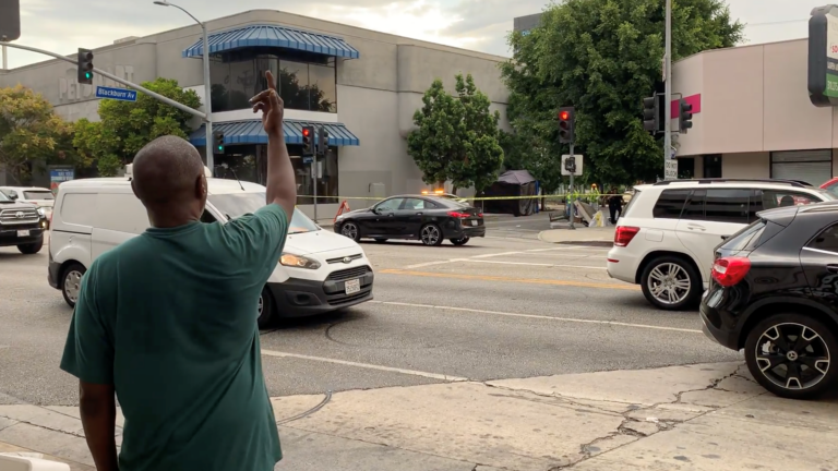 A man in a green shirt stands on one side of the street showing his middle finger to sanitation workers on the other side who are destroying his belongings.